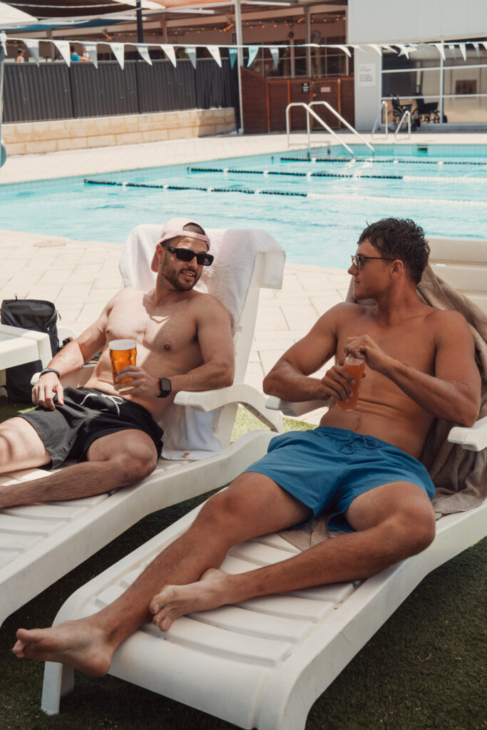 Two members relaxing on sun loungers beside the outdoor pool at ROAR Active Bibra Lake with drinks in hand.